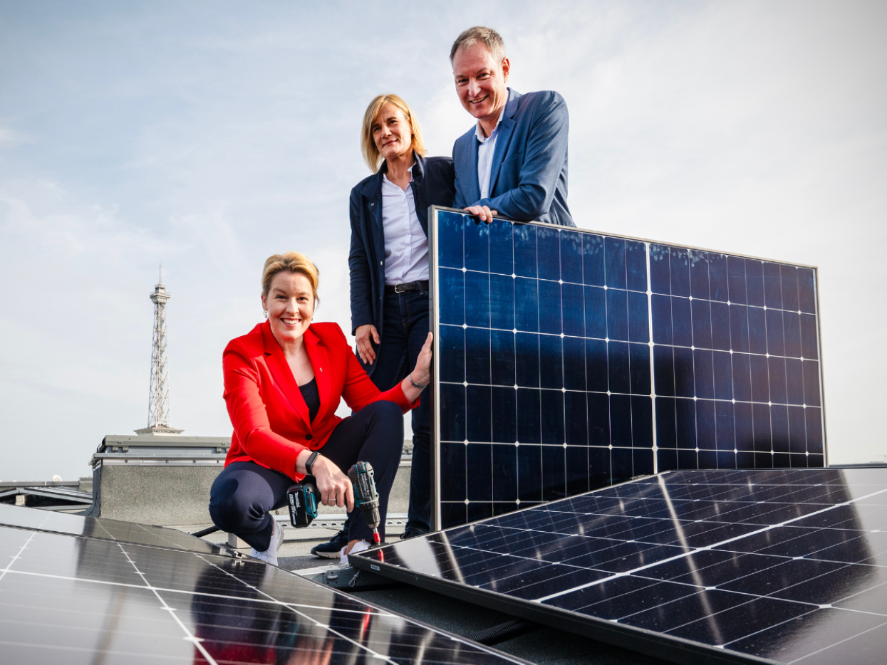 Mario Tobias steht mit Wirtschaftssenatorin Franziska Giffey und der Geschäftsführerin der Berliner Stadtwerke, Kerstin Busch vor einer Solarpanele auf einem Hallendach. Im Hintergrund der Funkturm.